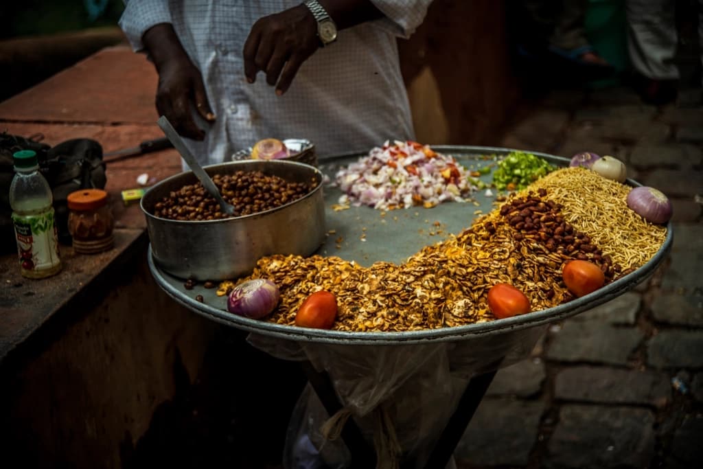 A street food vendor working over a large steel thali of chaat, spooning chana from a pot.